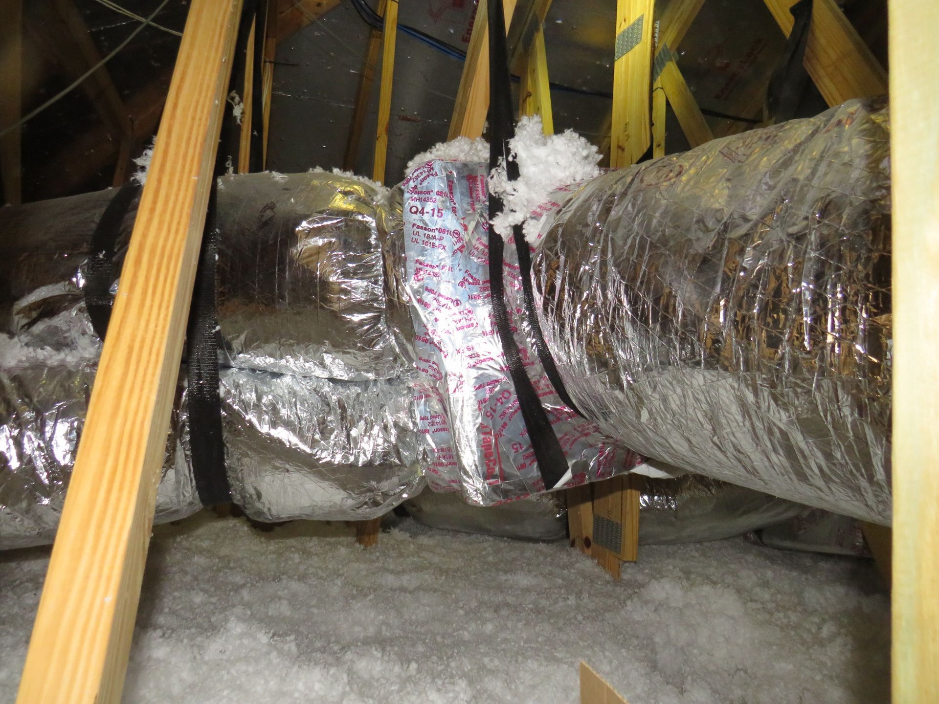Insulated flexible HVAC air ducts suspended with black straps in an attic filled with white loose-fill insulation.
