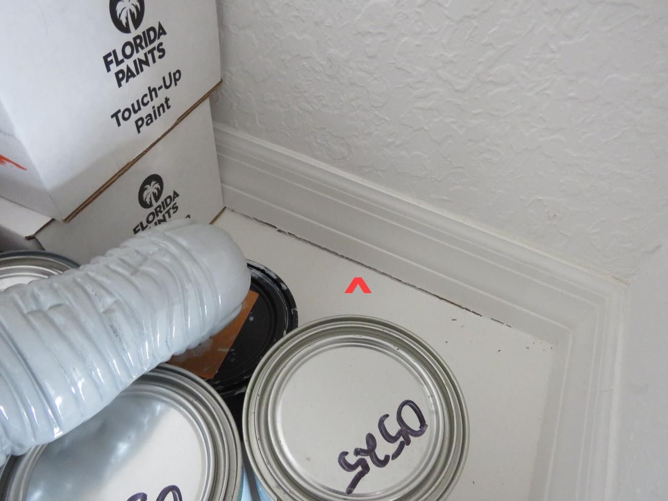 Several paint cans and a plastic bottle are stored on the floor against a white baseboard and wall corner.