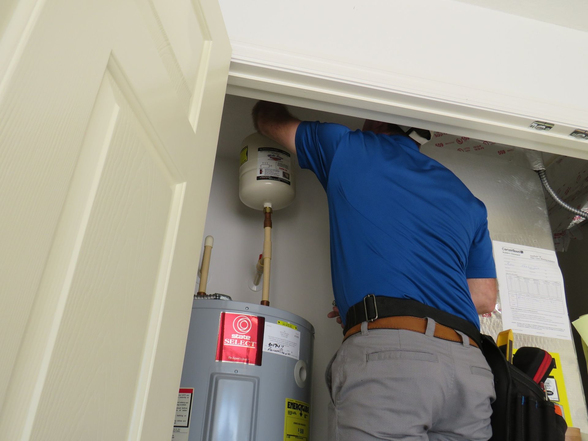 A technician in a blue shirt works on a water heater and expansion tank inside a utility closet.