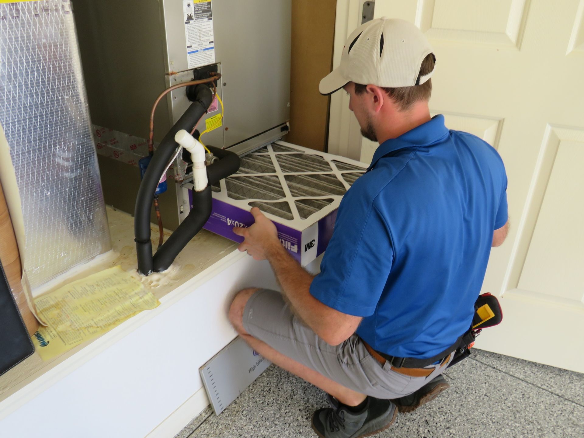 A technician in a blue shirt kneeling to replace an air filter in a residential HVAC unit.