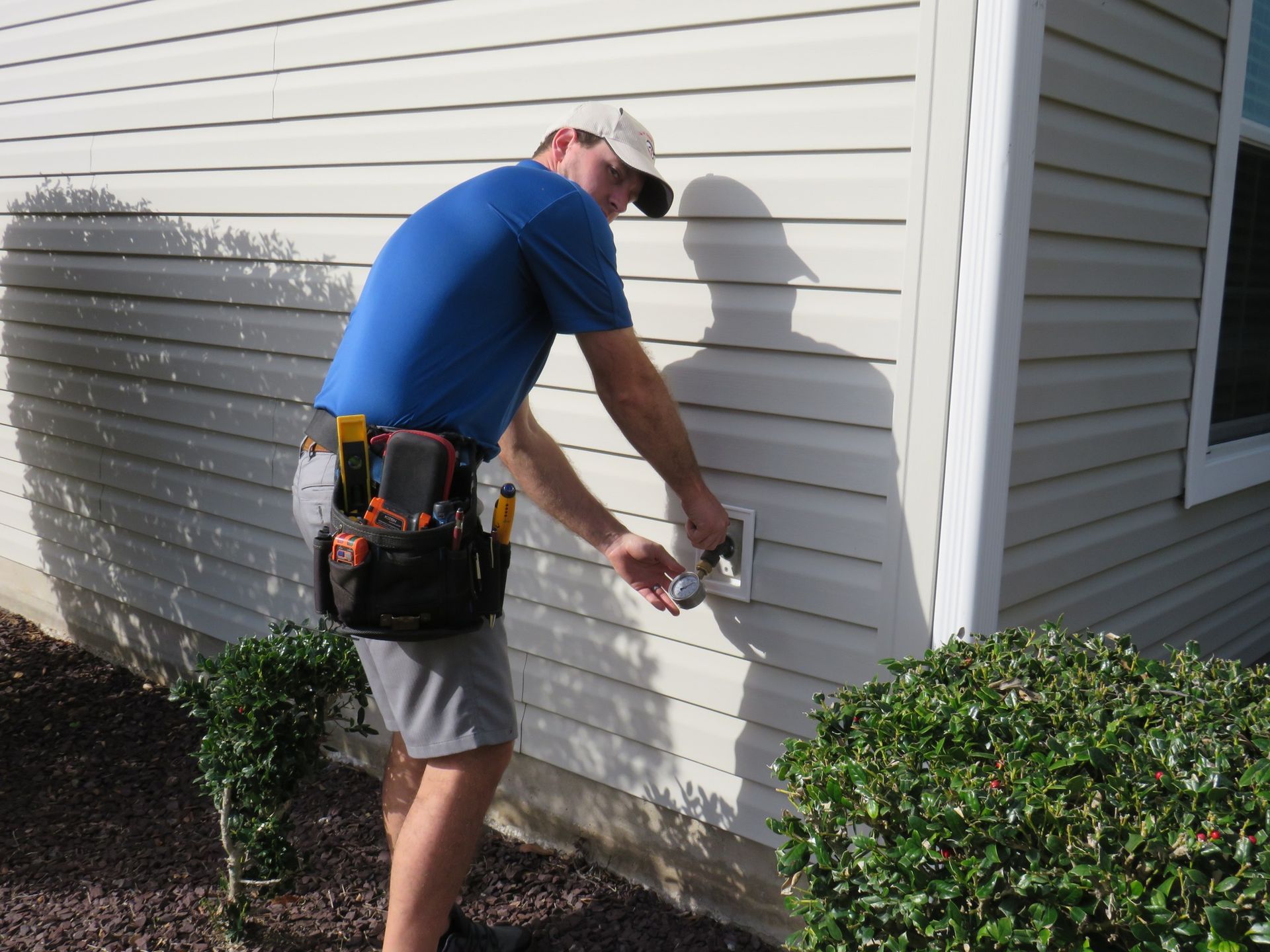 A service technician in a blue shirt and tool belt works on a utility connection mounted on a residential exterior wall.