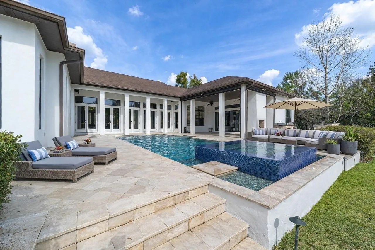 A sunlit patio features a swimming pool with a tiled hot tub, lounge chairs, and a white house under a blue sky.