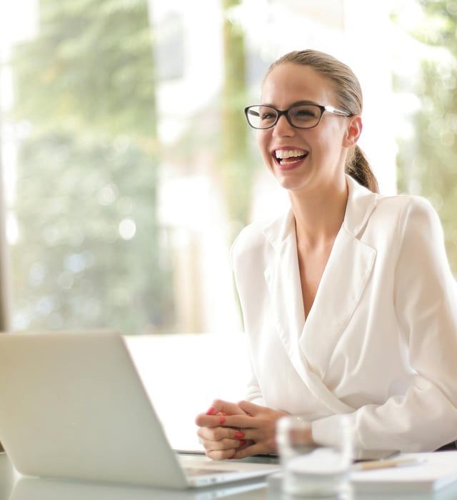 A woman wearing glasses is smiling while sitting in front of a laptop computer.