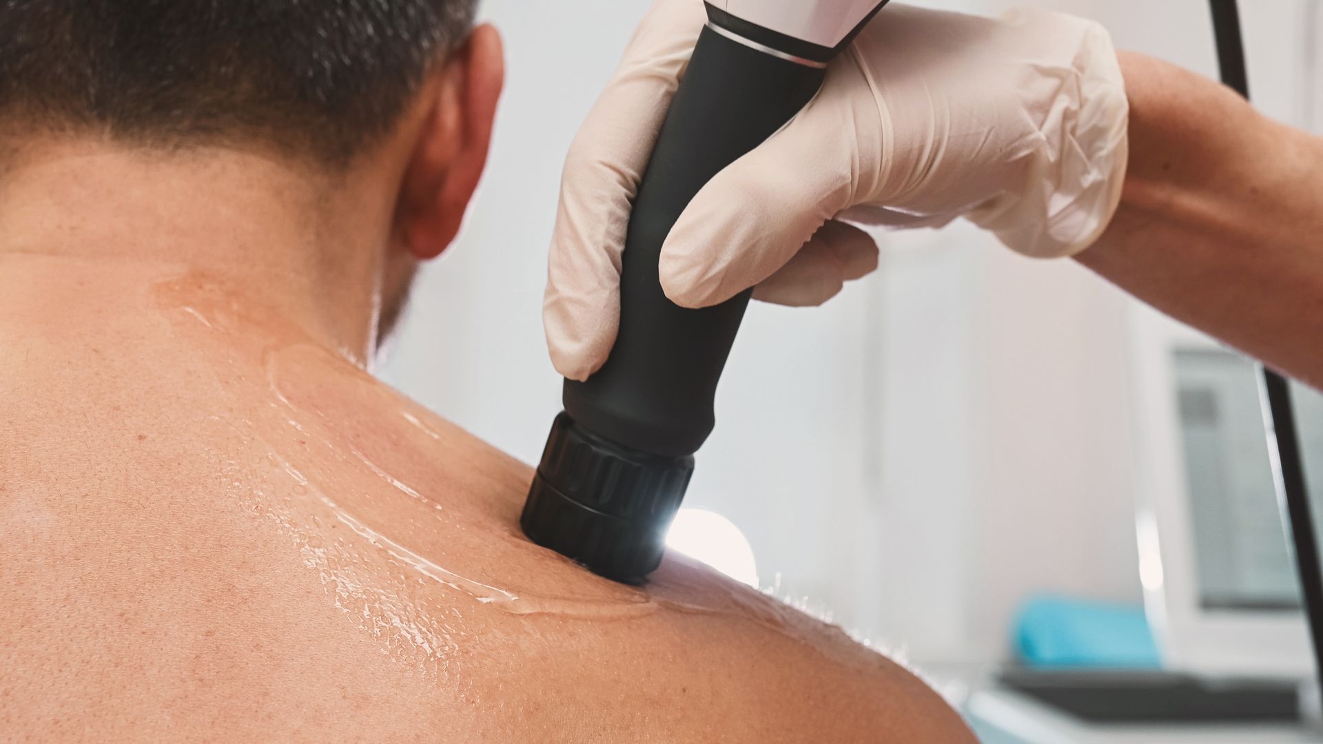 A practitioner in medical gloves uses an ultrasound therapy device on a person's shoulder covered in conductive gel.