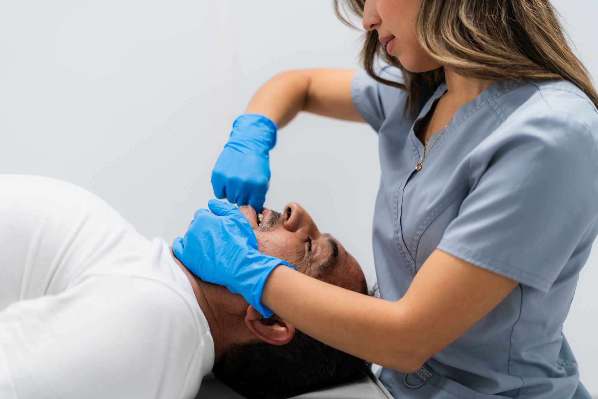 A healthcare worker in blue gloves performs an intraoral massage on a patient lying down in a clinical setting.