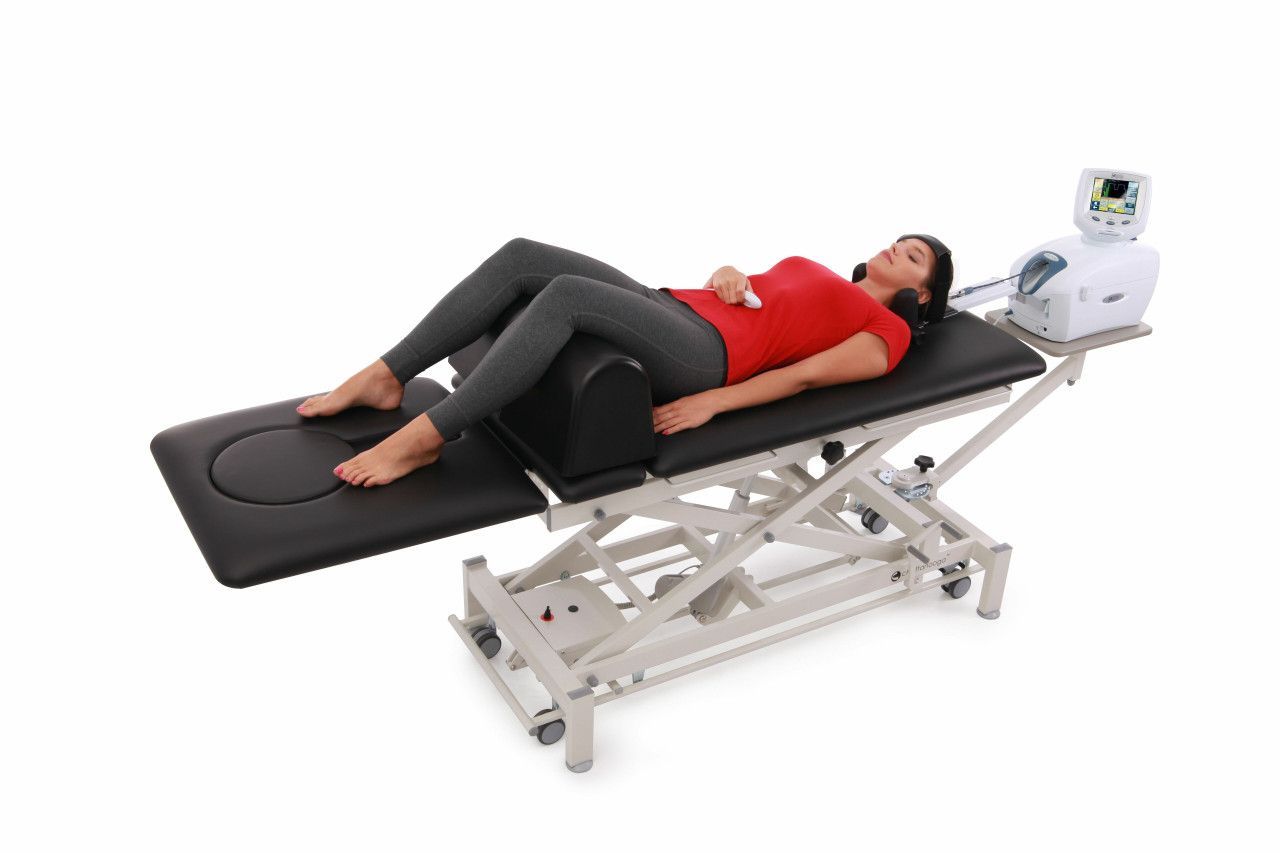 A person lies on a medical examination table with legs elevated on a bolster, near a clinical monitoring device.