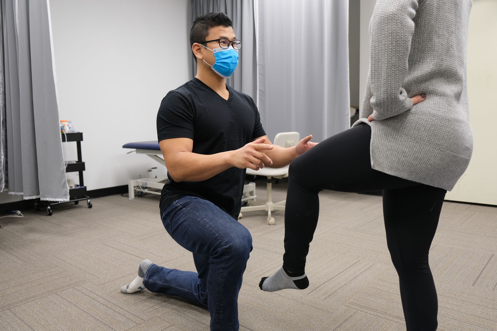 Physical therapist assisting a patient with a leg lift in a clinic. Both are masked.