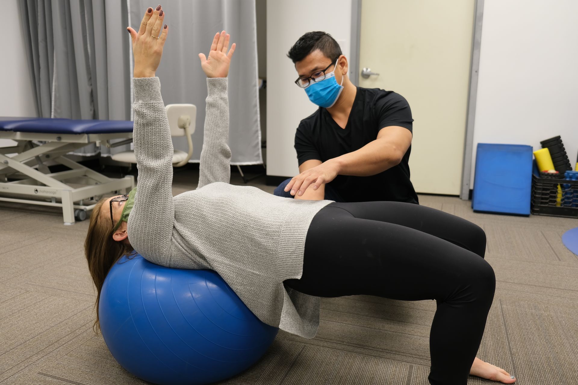 Woman on exercise ball with arms up, PT assisting, in a therapy setting.