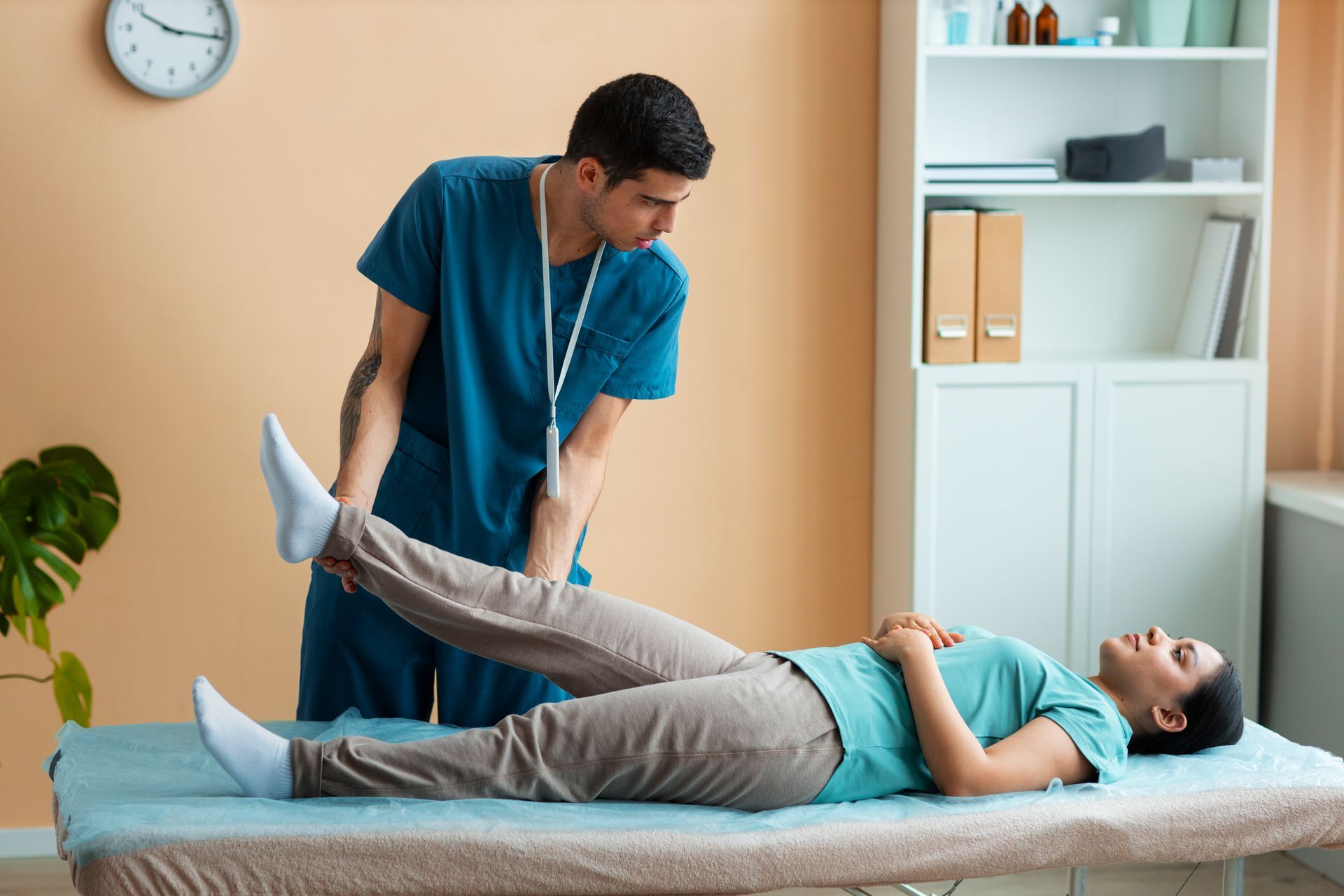 Physiotherapist examining woman's leg on a medical table; light-colored room, tan walls, teal scrubs.