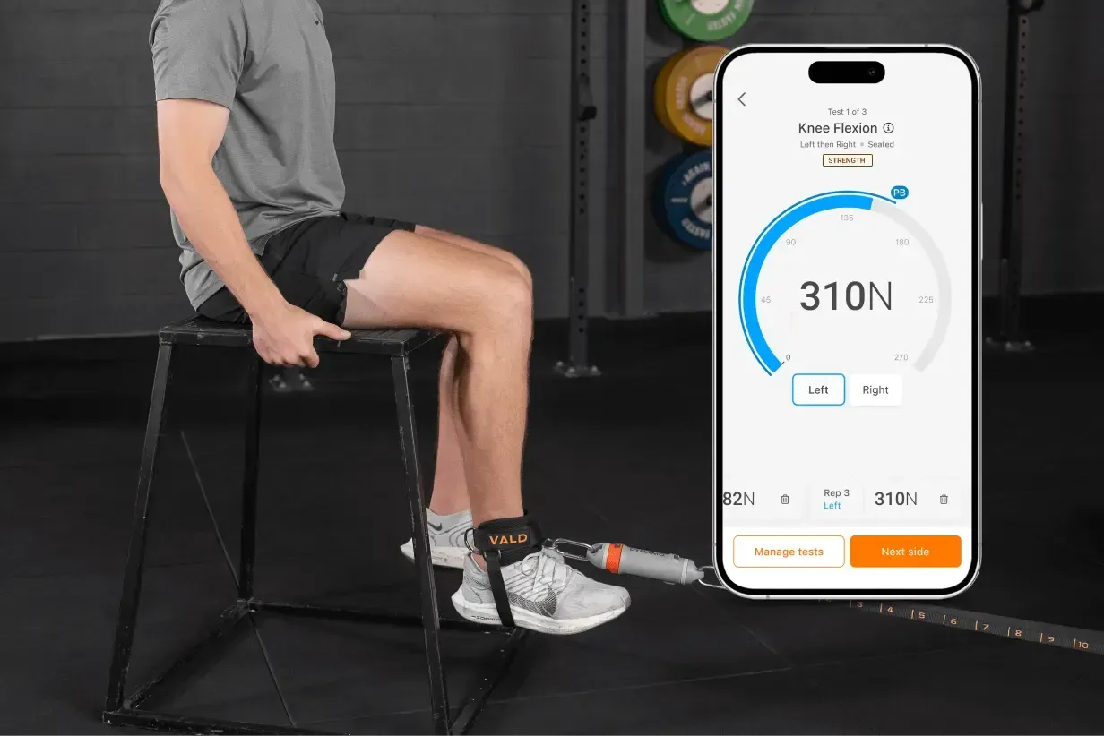 Man seated on a box, using a leg strength testing device connected to a smartphone displaying 310N of force.