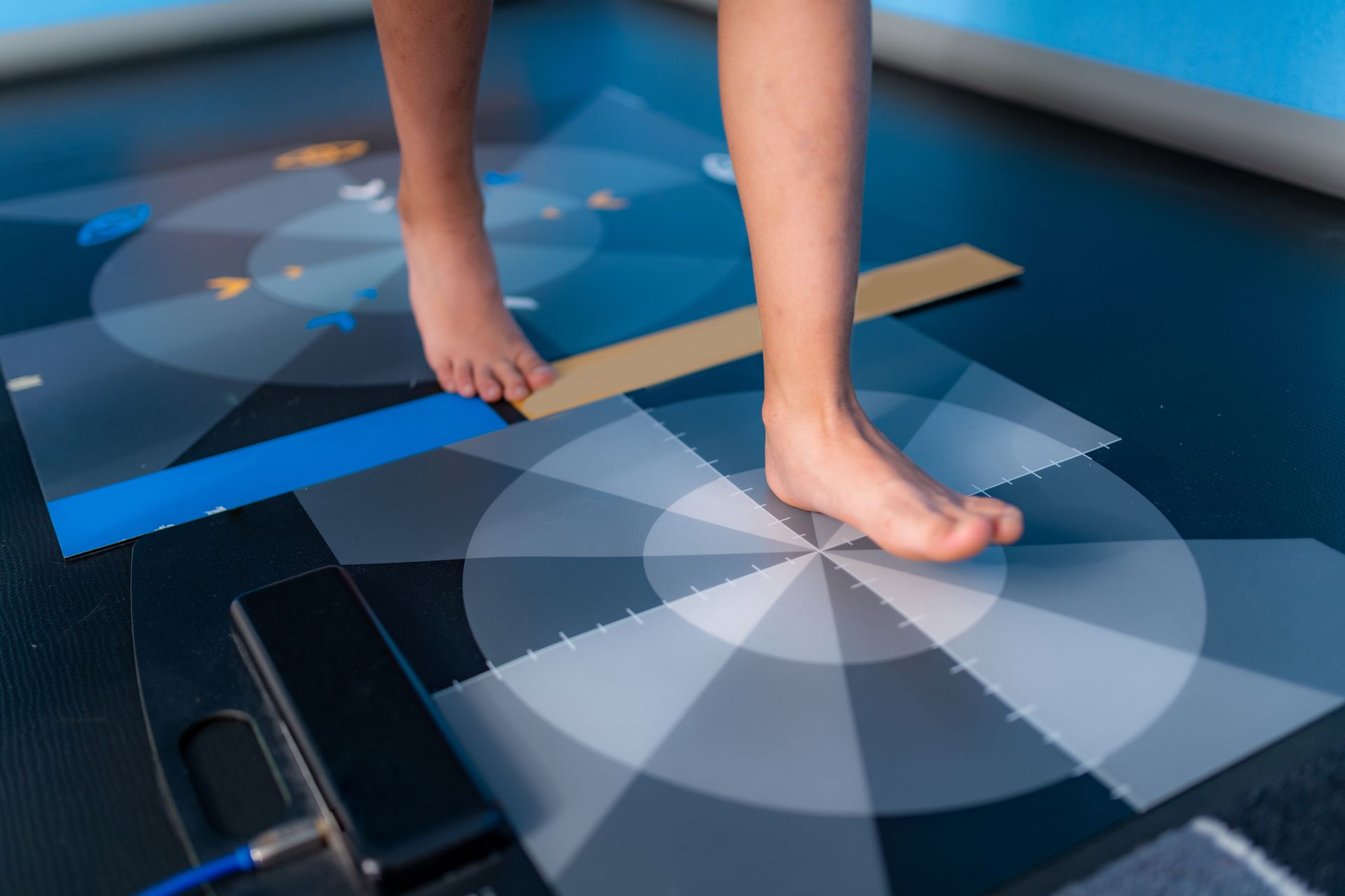 A person's bare feet on a mat with concentric circles and lines; a blue cable and black box are on the left.