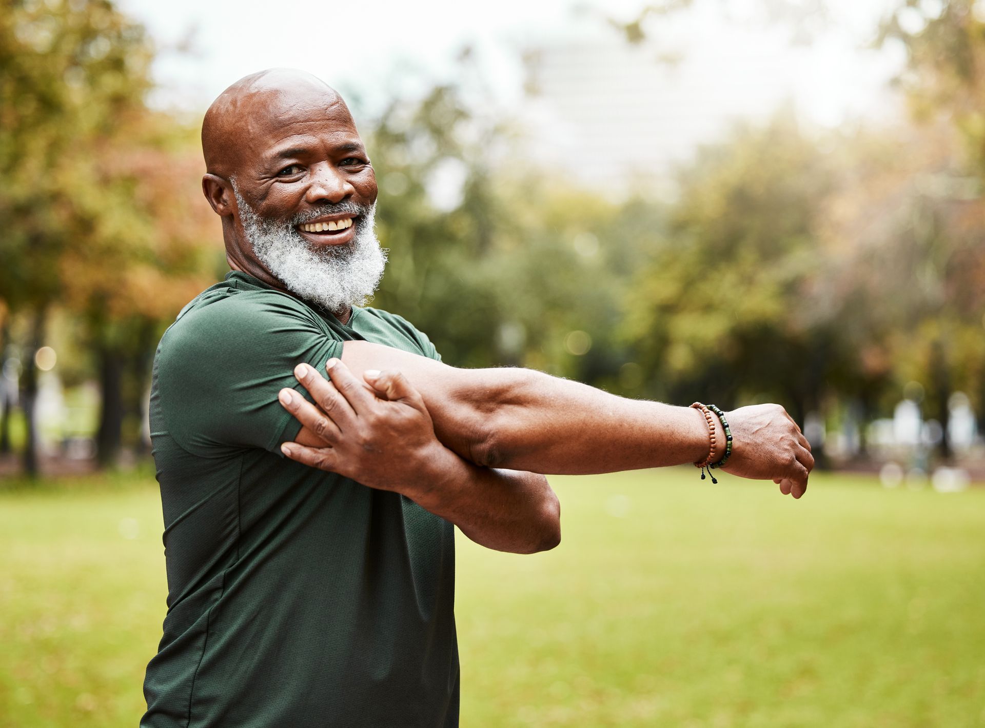 Smiling Black man in a green shirt stretches arm in a park, trees in the background.