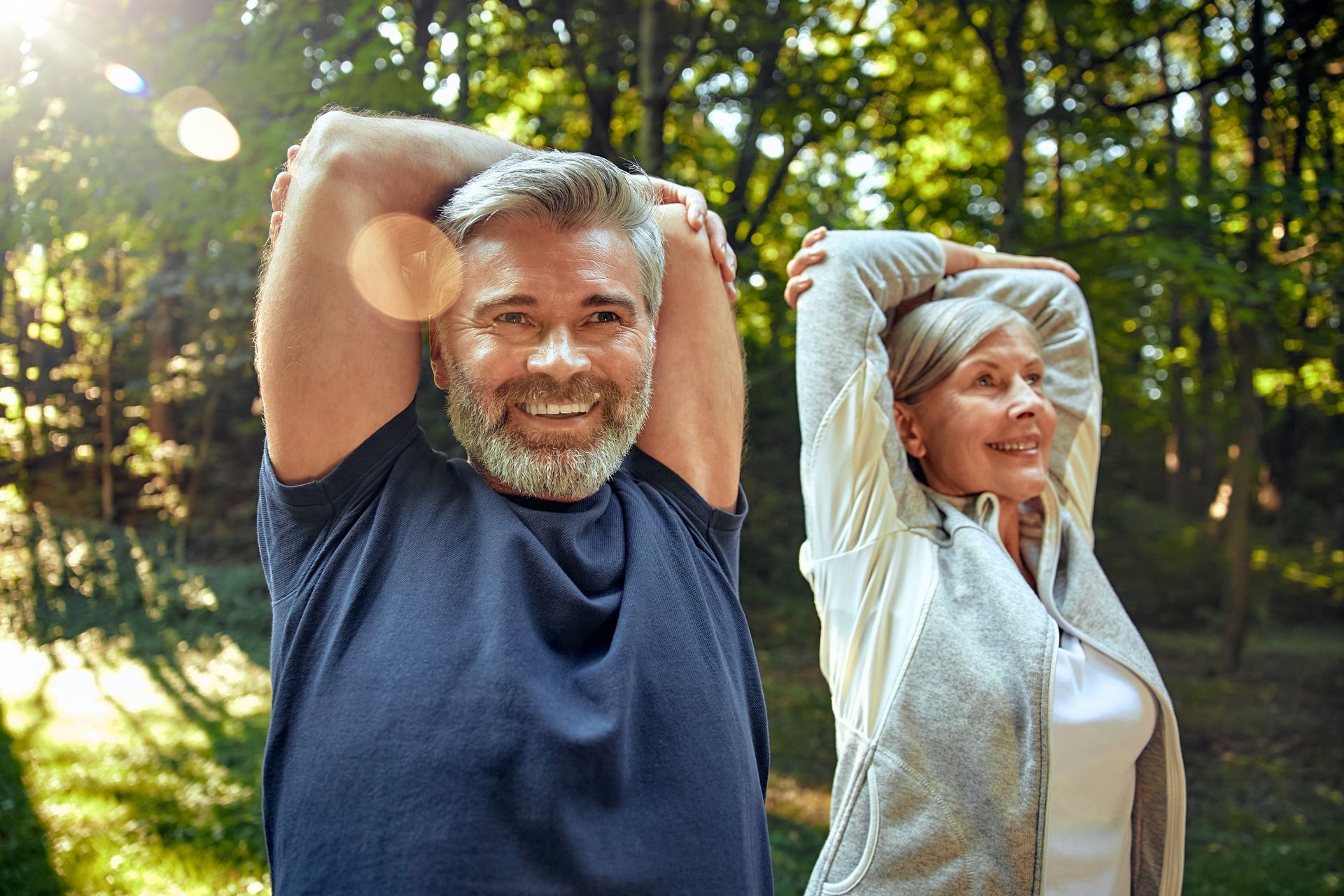 Mature couple stretching arms in a sunlit forest, smiling.