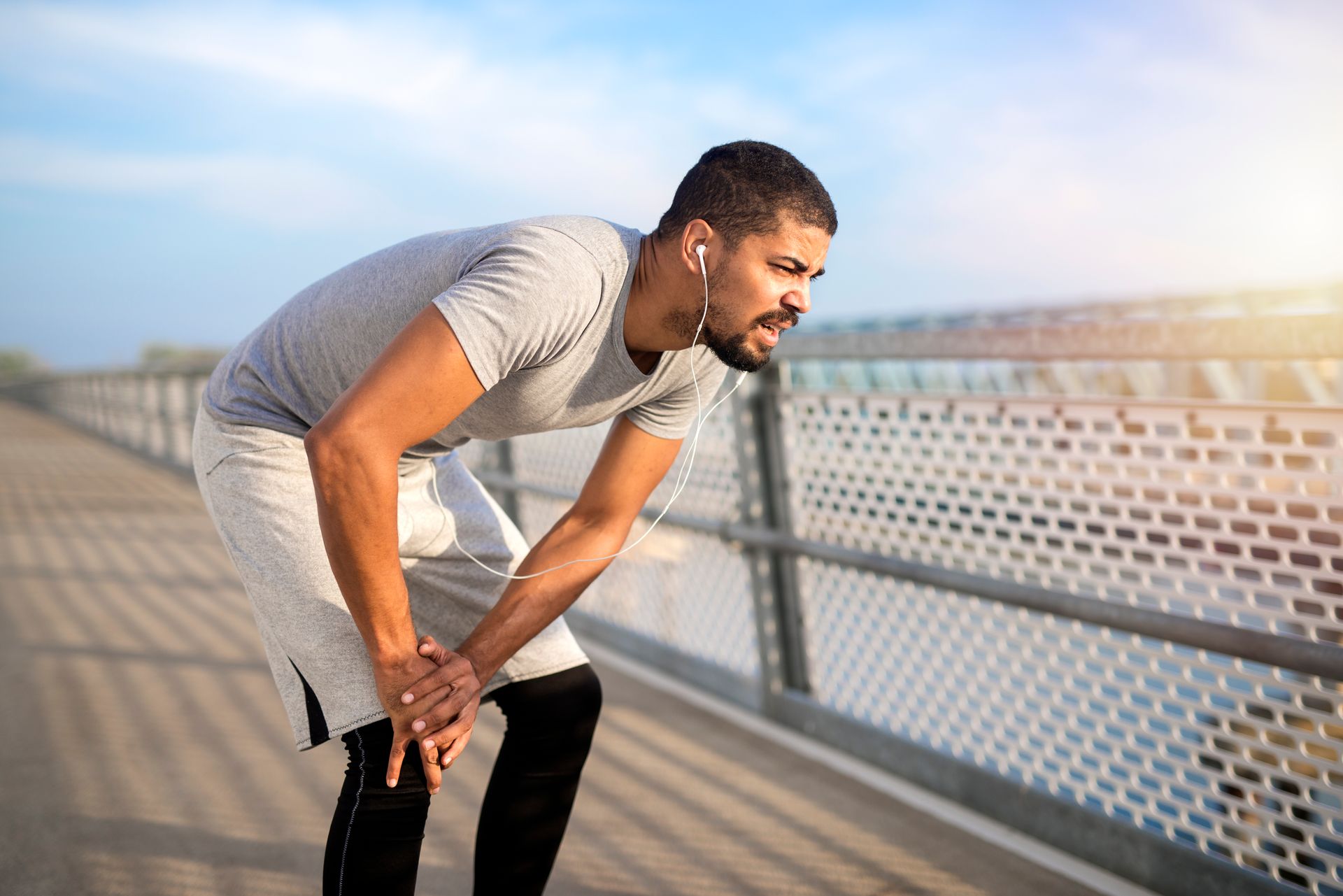 Man in gray shirt and shorts, resting on a bridge, holding his knee, appears in pain.