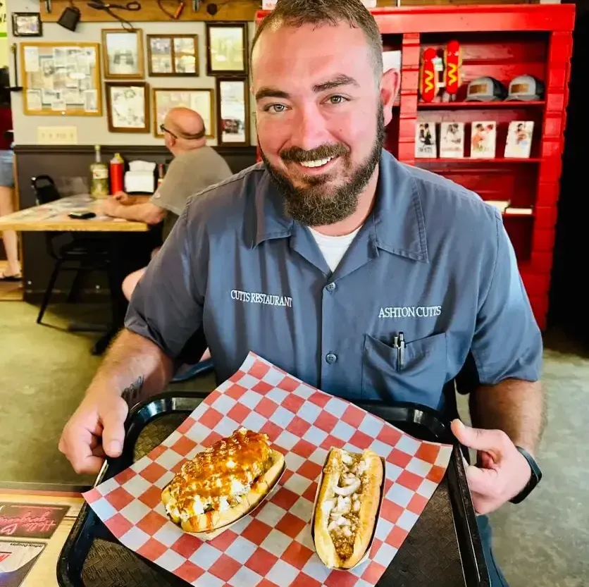 Man smiling, holding a tray with two hot dogs, inside a restaurant. He's wearing a gray shirt; the setting is brightly lit.