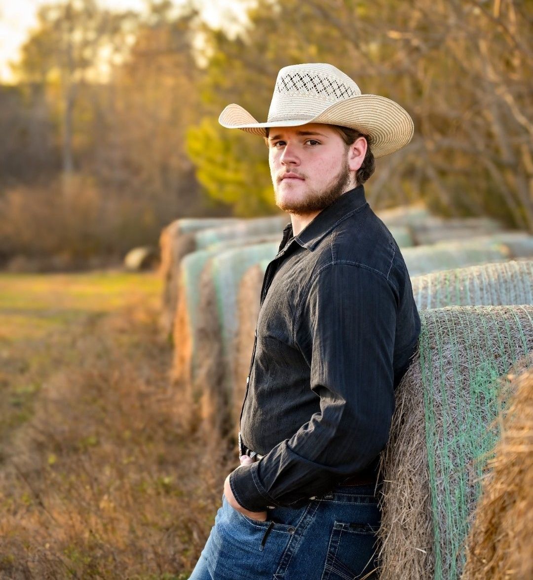 A man wearing a cowboy hat is leaning against a hay bale.