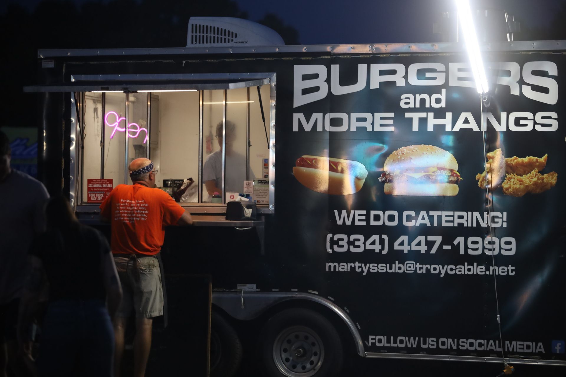 A man standing in front of a food truck that says burgers and more thangs