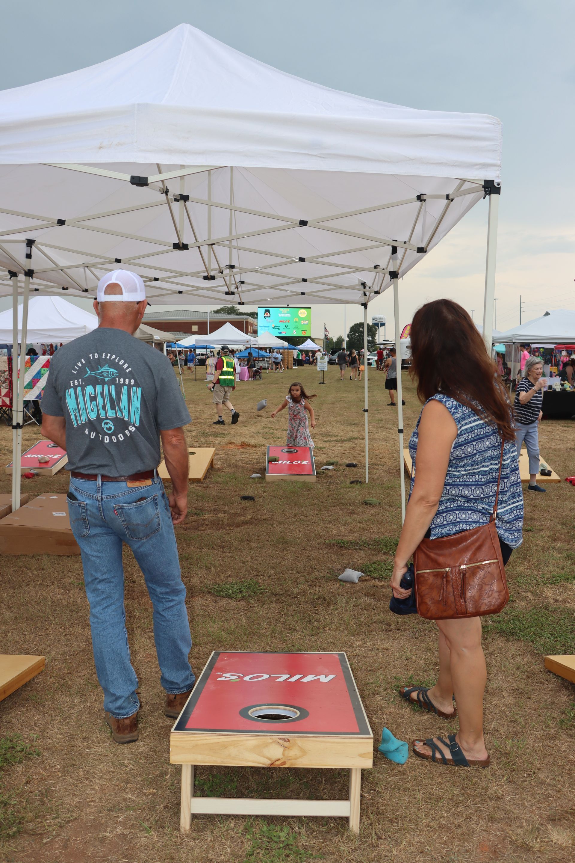 People playing cornhole at an outdoor event. A man and woman stand near a red and black cornhole board as others play in the distance.