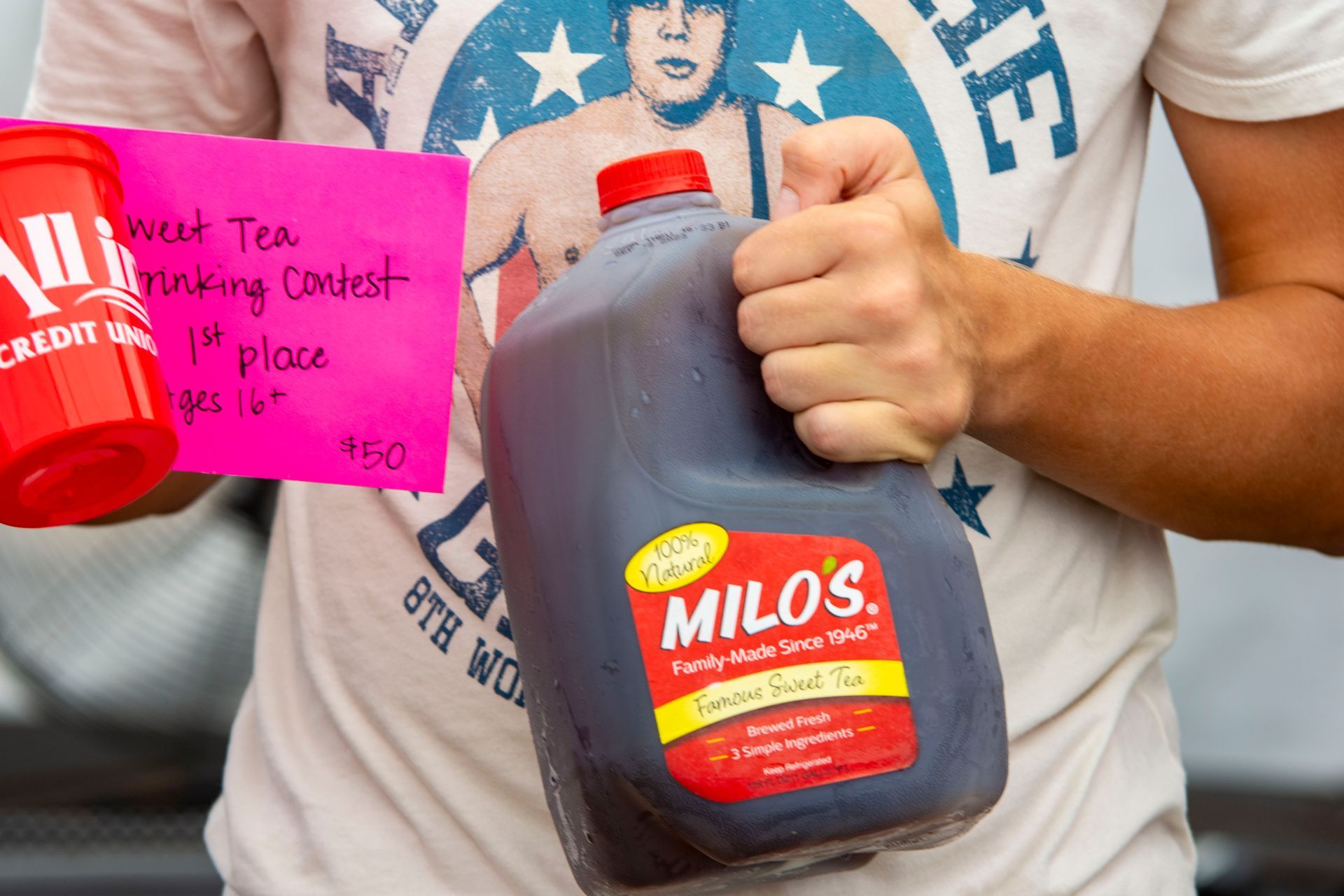 A person holding a large jug of Milo's Famous Sweet Tea.  They're also holding a pink note that says 