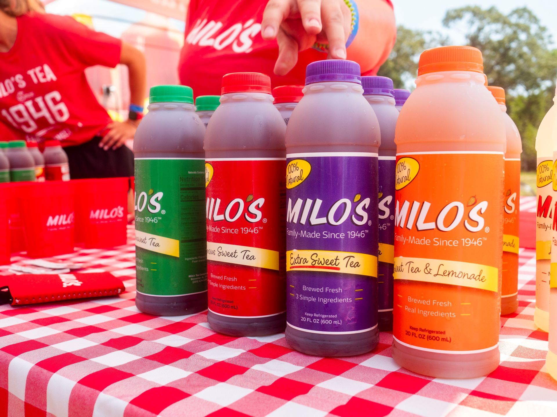Bottles of Milo's tea in various flavors on a checkered table. People in the background, likely at an event.
