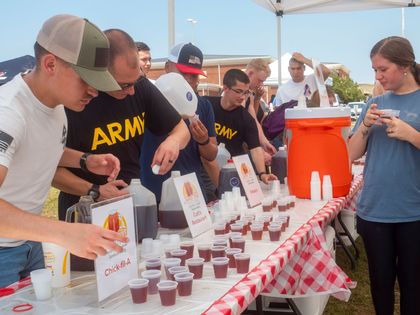 A group of people are standing around a table with a sign that says army on it. Sweet Tea tasting contest