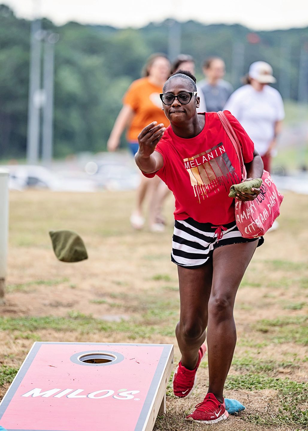 Woman in red shirt and striped shorts throws a beanbag at a cornhole board outdoors.