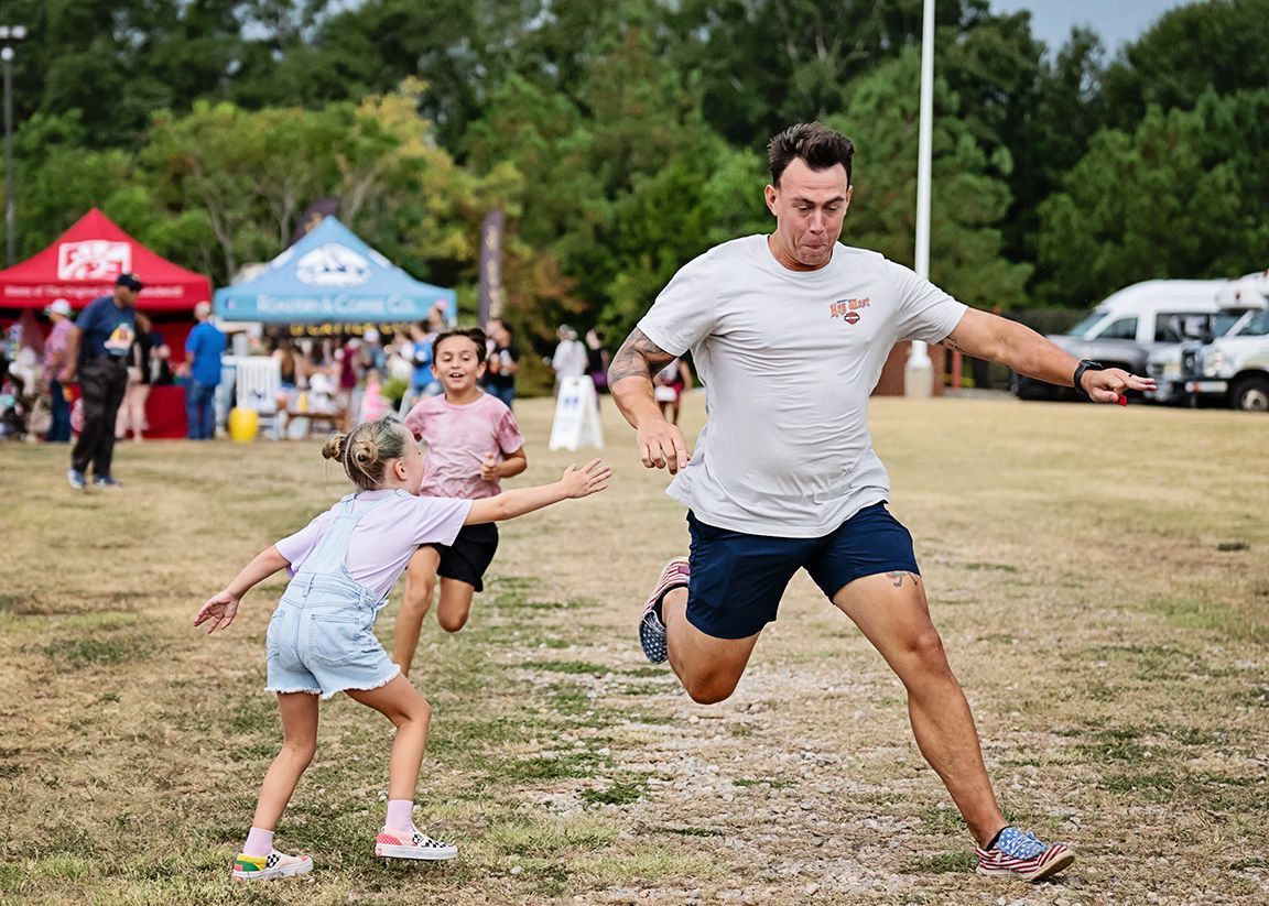 A man in shorts and a t-shirt runs in a field, chased by two children near tents and trees.