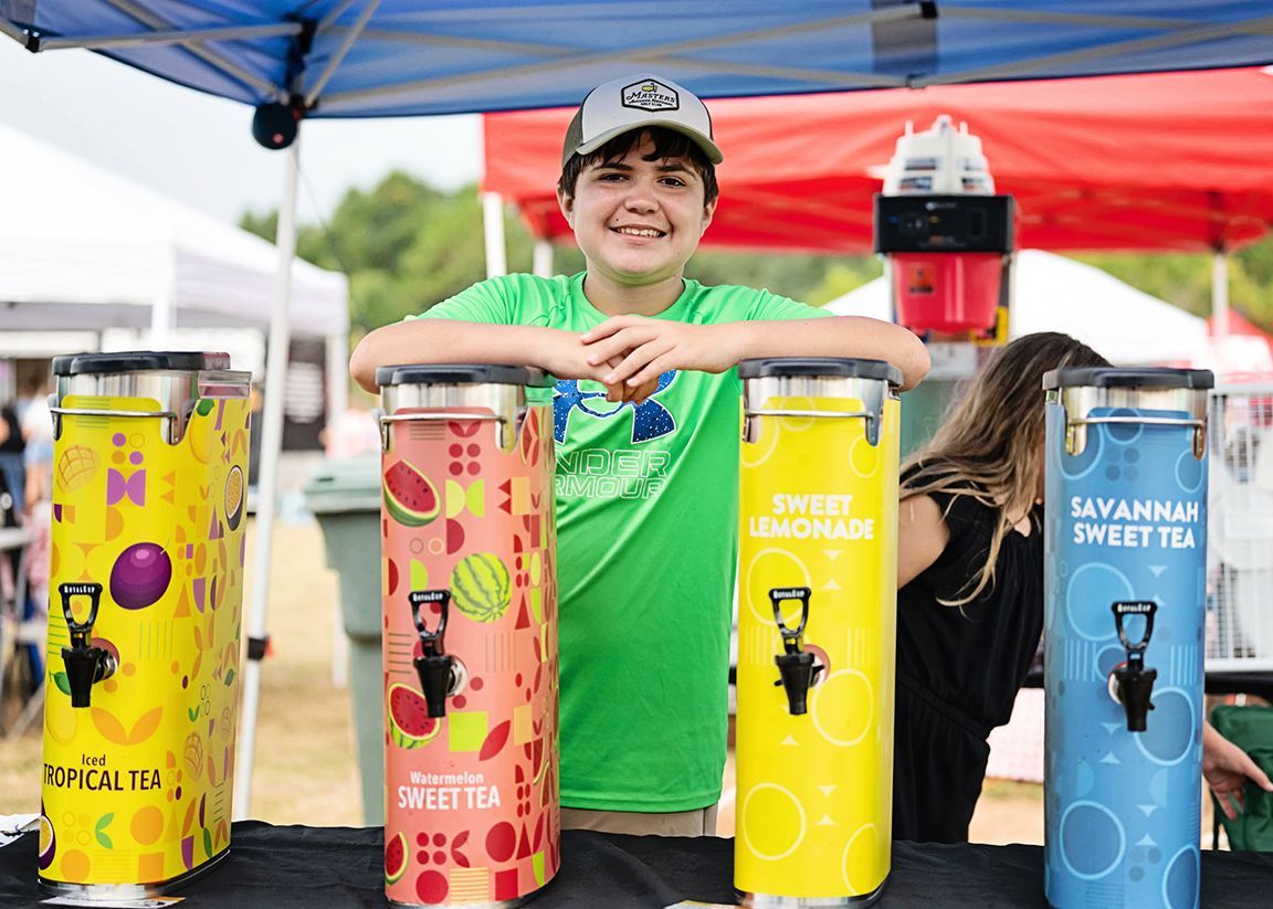 A young boy is standing behind a table with drink dispensers.