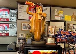 A whimsical statue of a hot dog person pouring ketchup and drinking a soda in a diner setting.