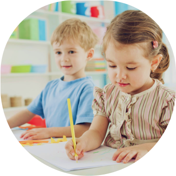 A young girl and boy are drawing at a table in a brightly colored classroom. The girl is focused on her work.