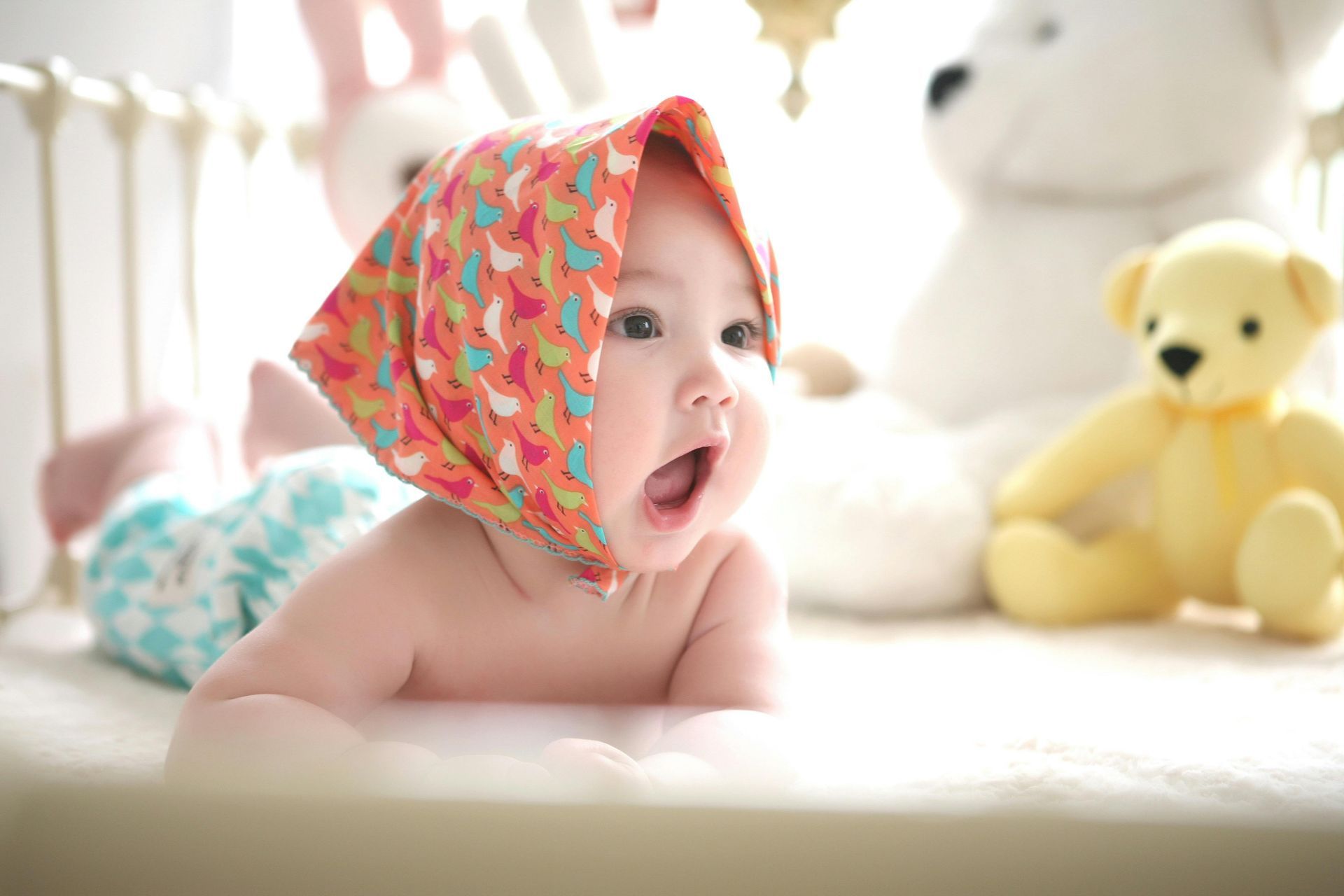 A baby wearing a head scarf is laying on its stomach on a bed.