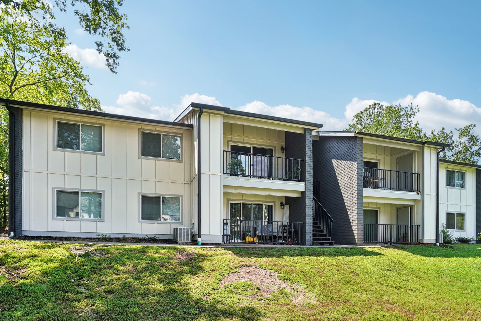Two-story apartment building with white siding, black trim, and balconies, on a grassy hill under a blue sky.