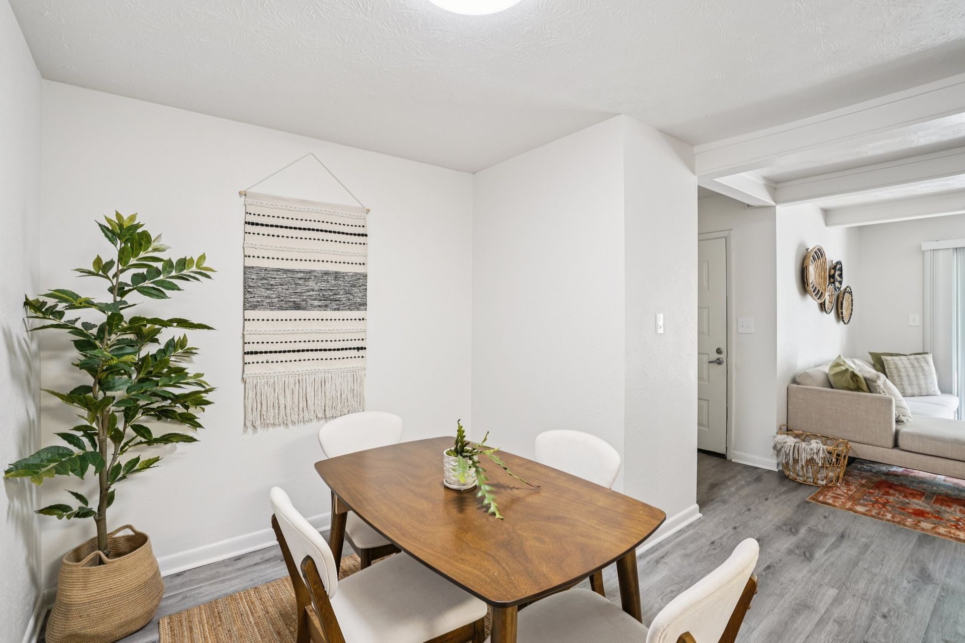 Dining area with wooden table, four white chairs, woven wall hanging, and a potted plant. Light gray floors and white walls.