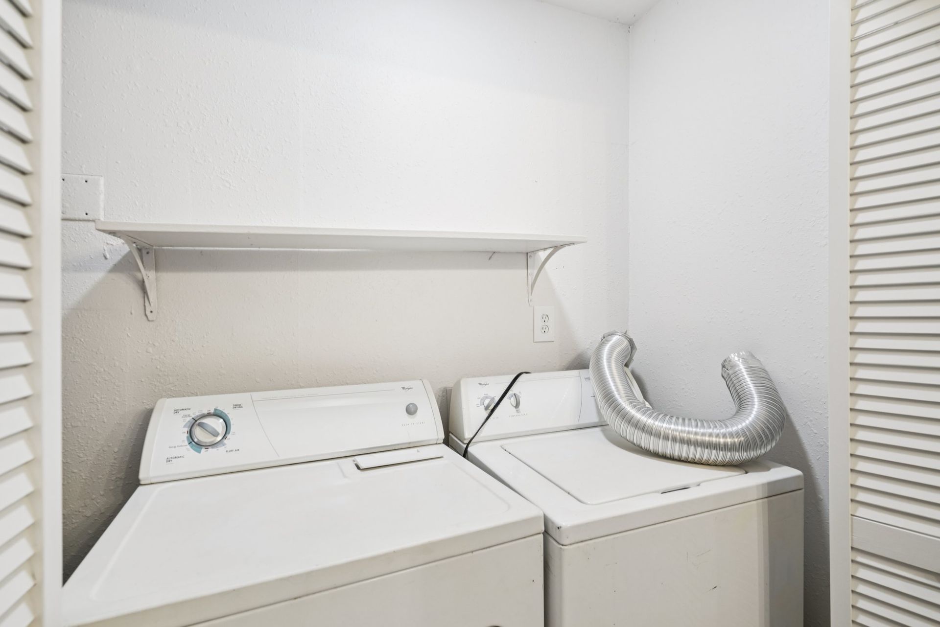 A white laundry room with a washer and dryer, shelf, and vent hose. The room is mostly white.