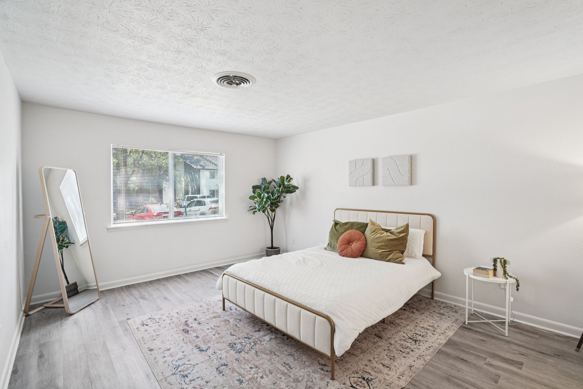 Bedroom with a bed, full-length mirror, small side table, and window. The walls are white, and the floor is light gray.