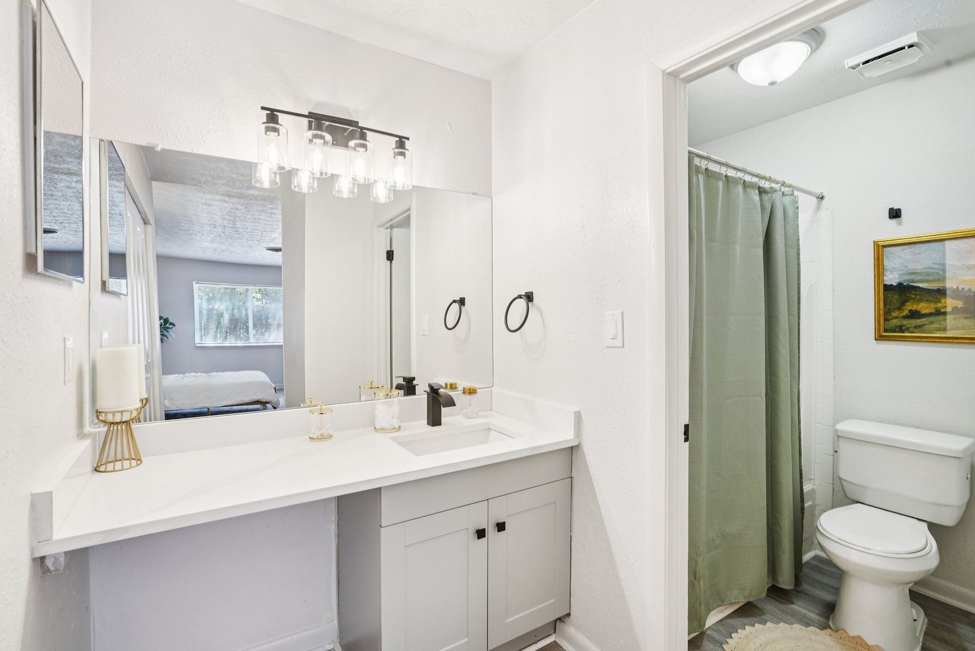 A newly renovated bathroom with a white vanity, black faucet, and a light green shower curtain. A mirror reflects a glimpse of a bedroom.