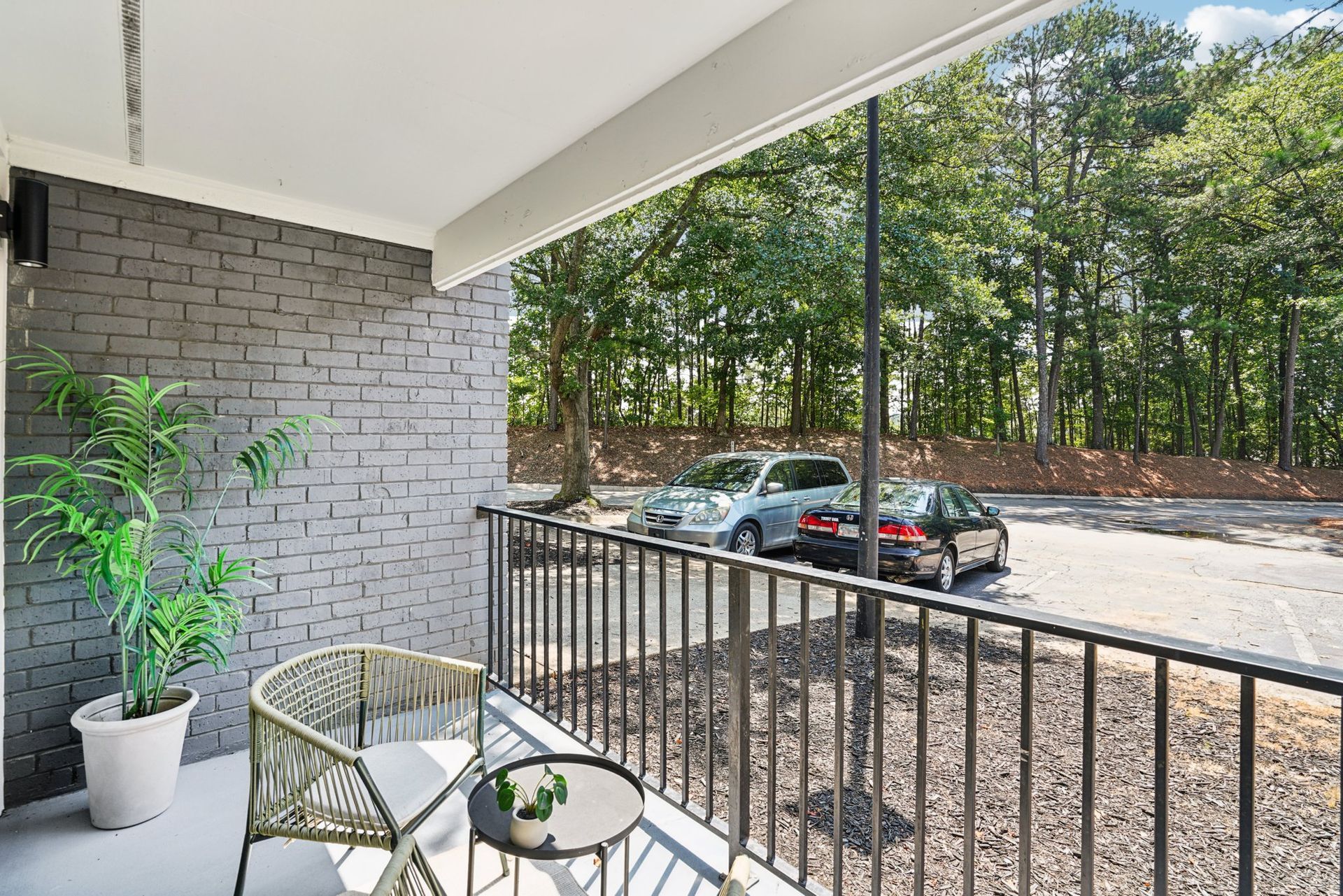 A balcony with a small table and chair faces a parking area with cars and a wooded area beyond.