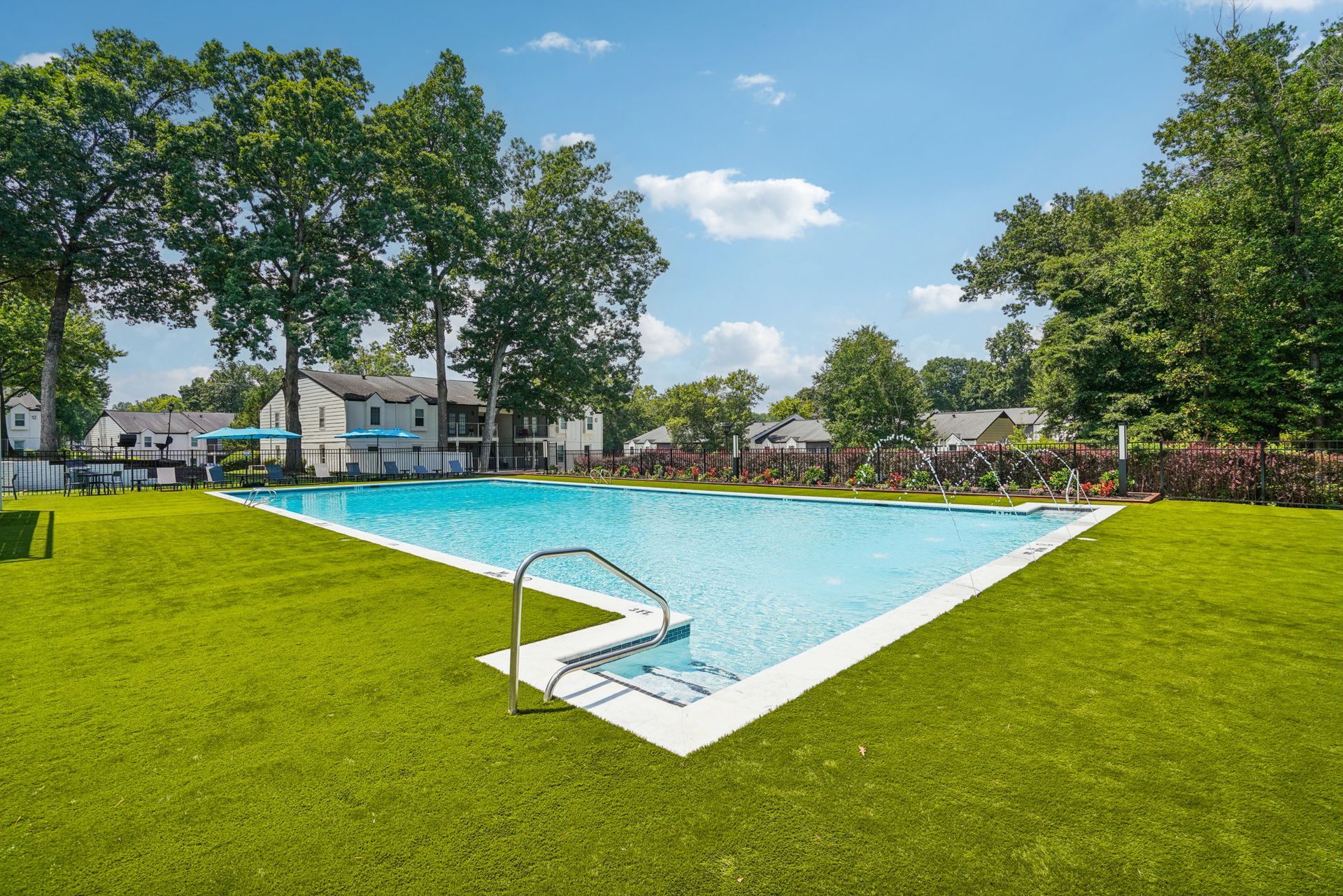 A rectangular swimming pool with a white border, surrounded by green grass and trees, under a blue sky.