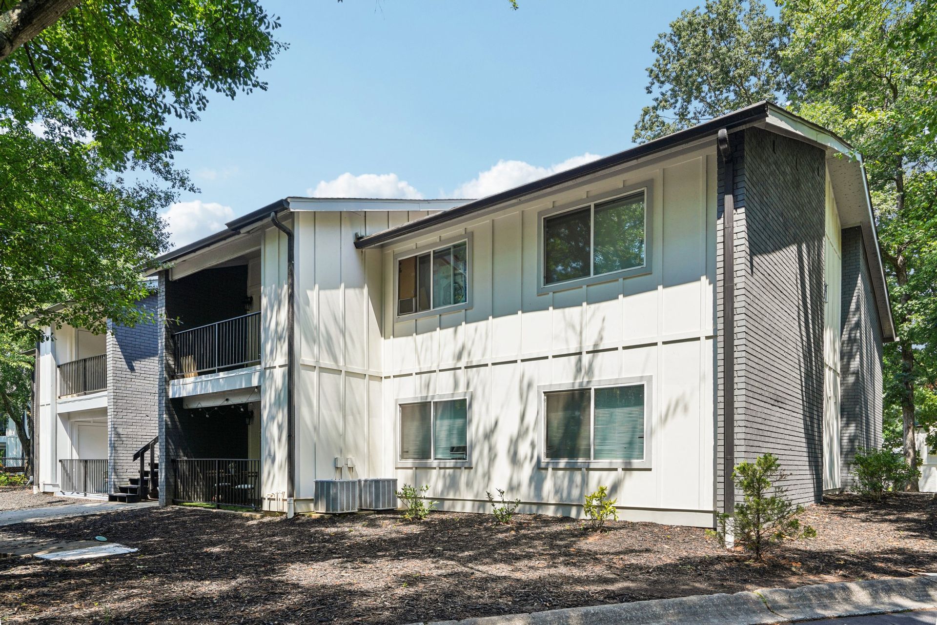 Two-story apartment building with white siding, a dark roof, and surrounding trees on a sunny day.
