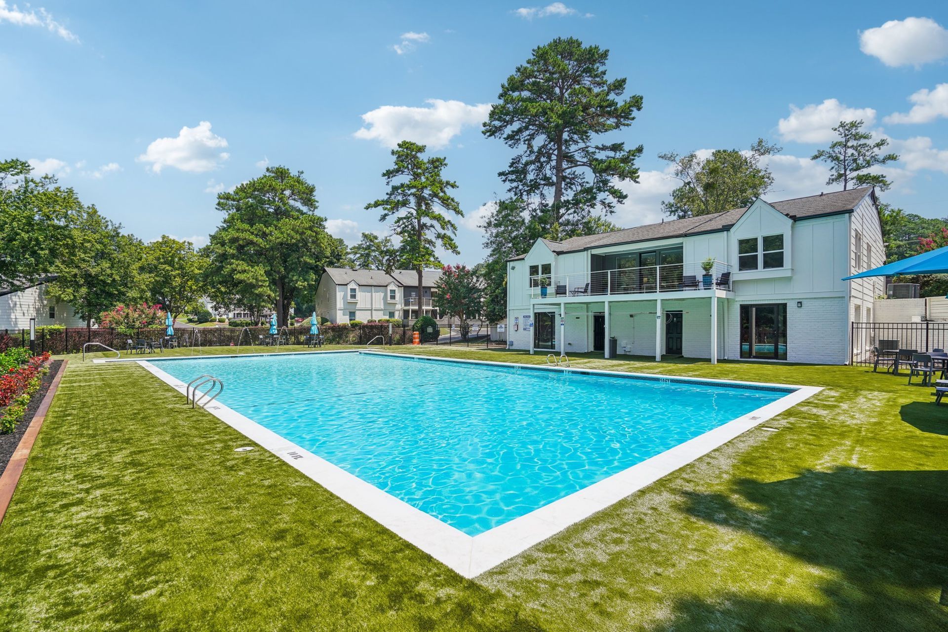 Swimming pool surrounded by green grass and white buildings. Blue sky with some trees in the background.