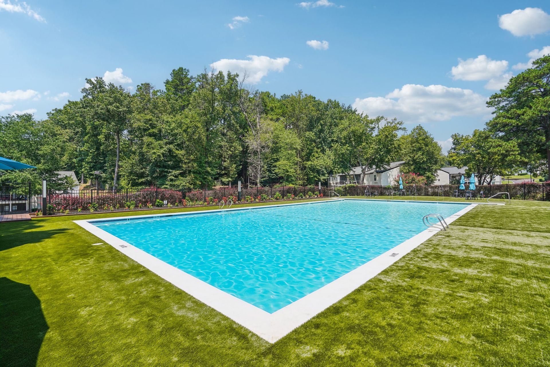 Swimming pool filled with blue water, surrounded by green grass, trees, and a clear blue sky.