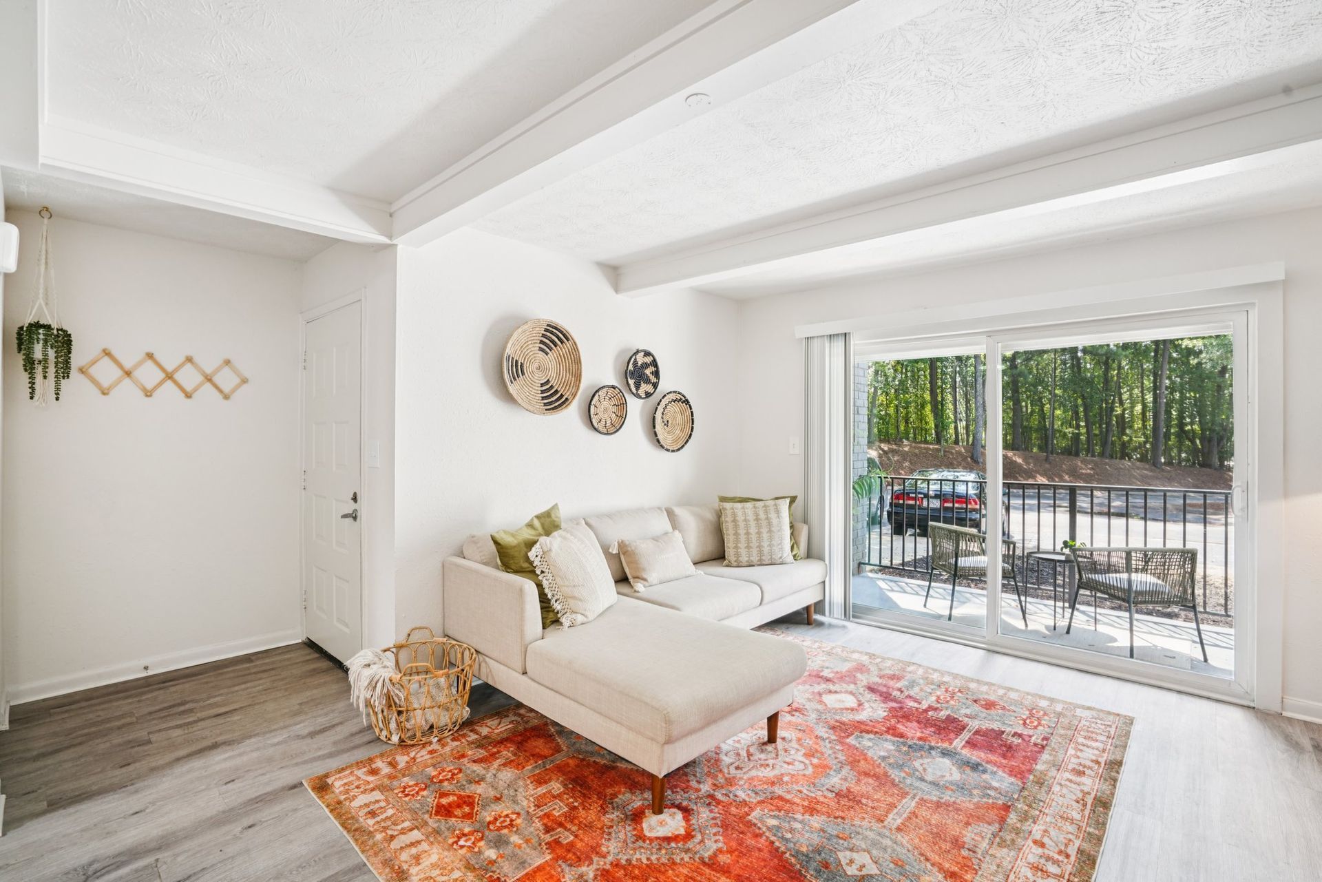 A bright living room with a white sofa and rug, opening to a balcony. Decor includes woven wall art and a coat rack.