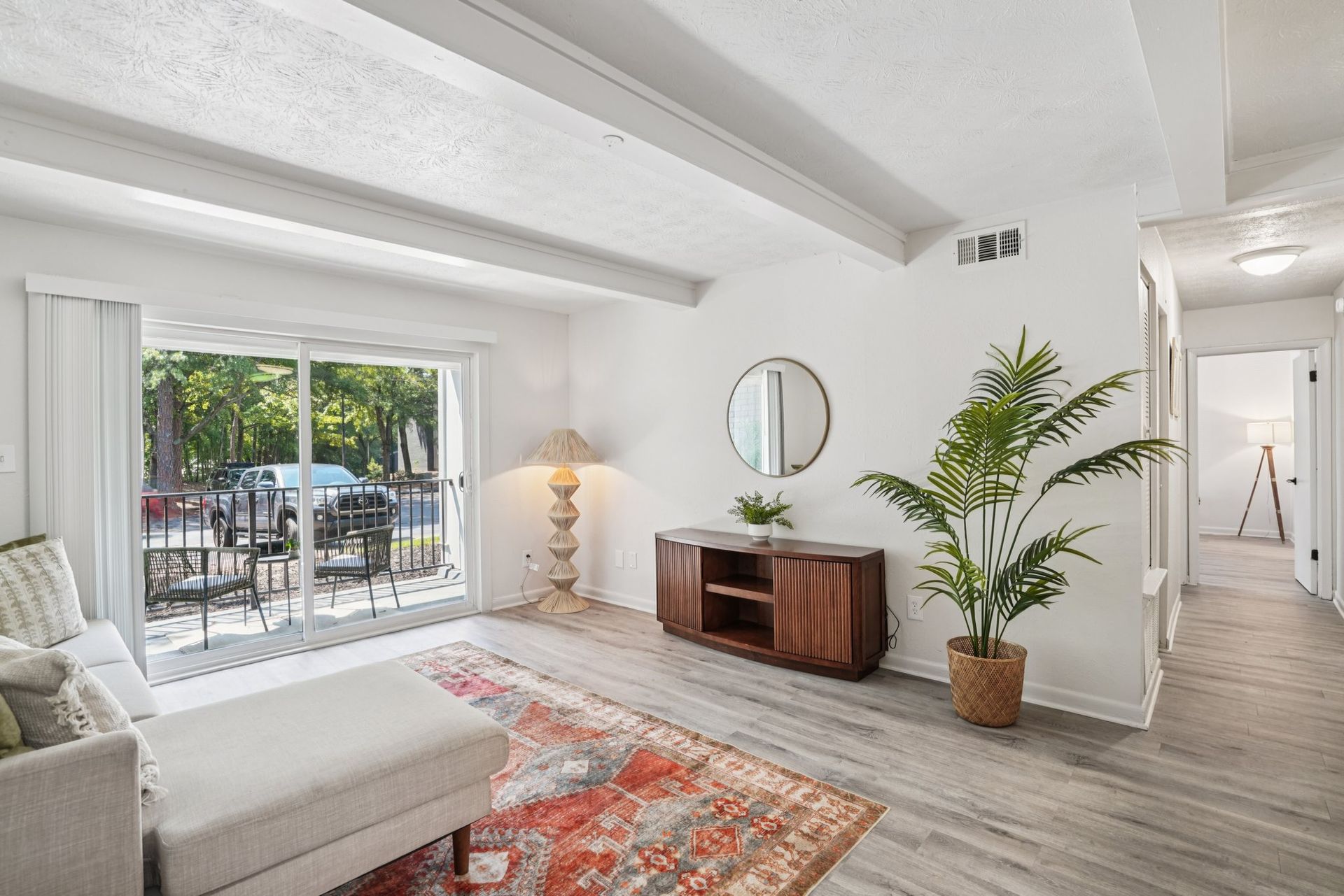 Bright living room with sliding glass doors, a beige sofa, and a wooden cabinet topped with a round mirror and a potted plant.