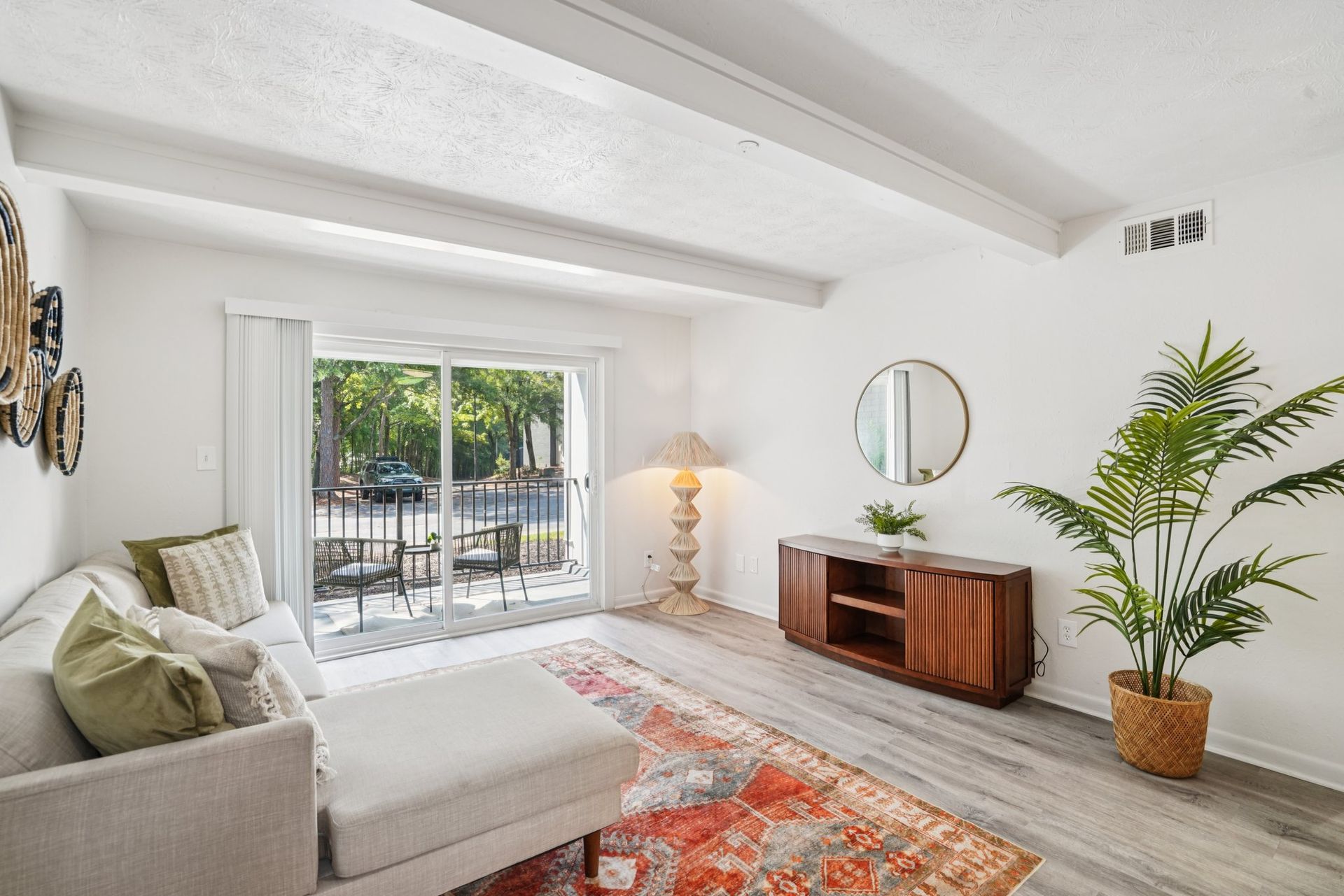 Living room with a white couch, wooden cabinet, and large window overlooking a patio. Warm rug and large potted plant.