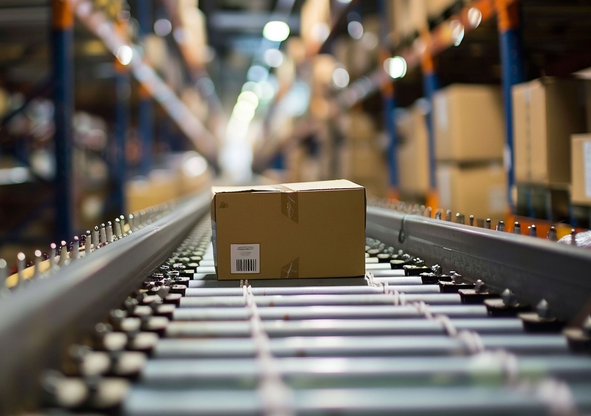 Cardboard box on a conveyor belt in a warehouse, moving towards a blurry background of shelves.