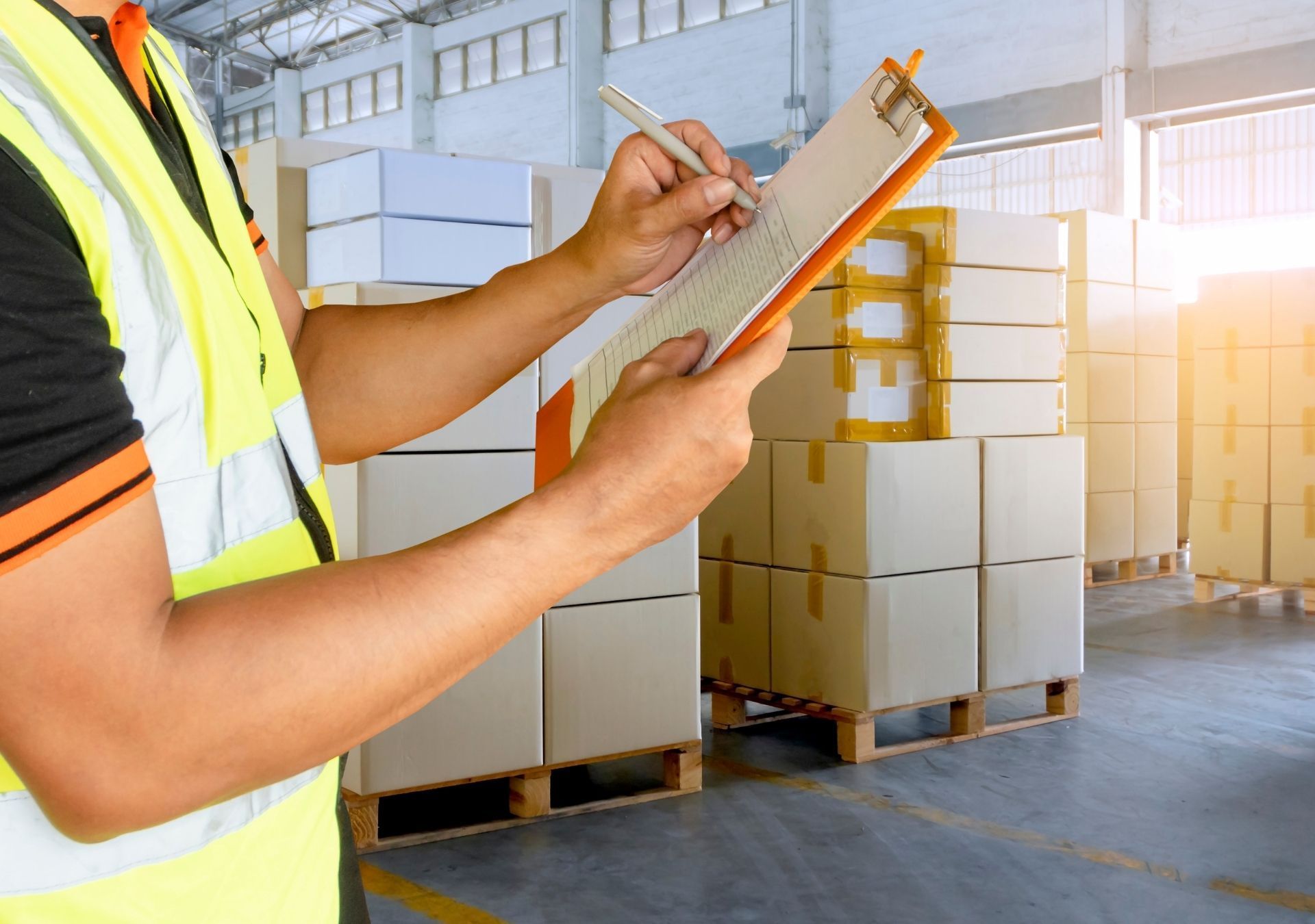 Warehouse worker in a safety vest writing on a clipboard, surrounded by stacked boxes.
