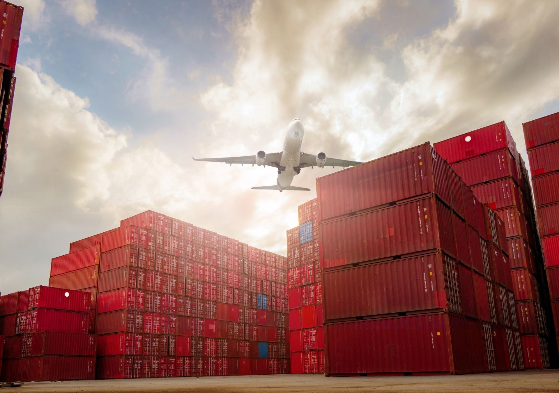 Airplane flying over red shipping containers at a port under a cloudy sky.