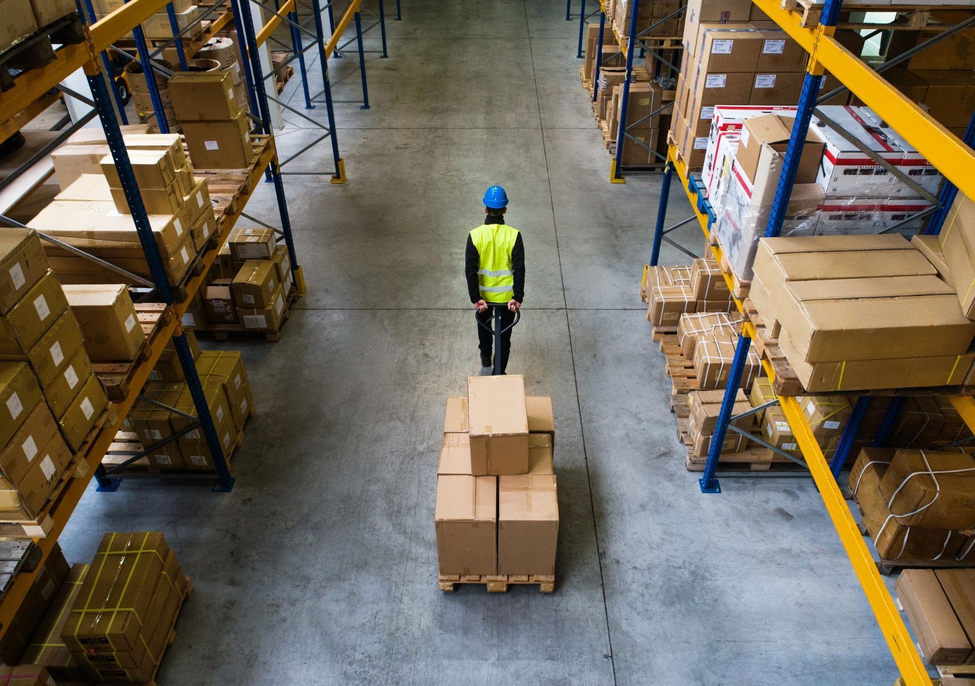 Warehouse worker on a pallet of boxes in an aisle surrounded by shelves of boxes.