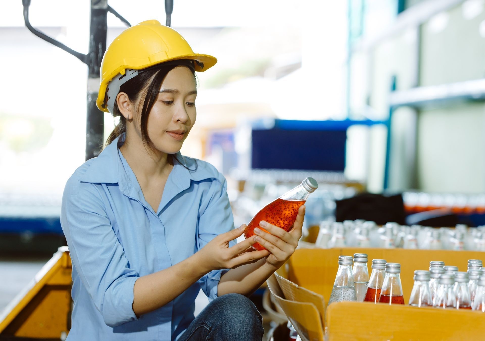 Woman in hard hat examines a bottle of red liquid in a warehouse, surrounded by bottled drinks.