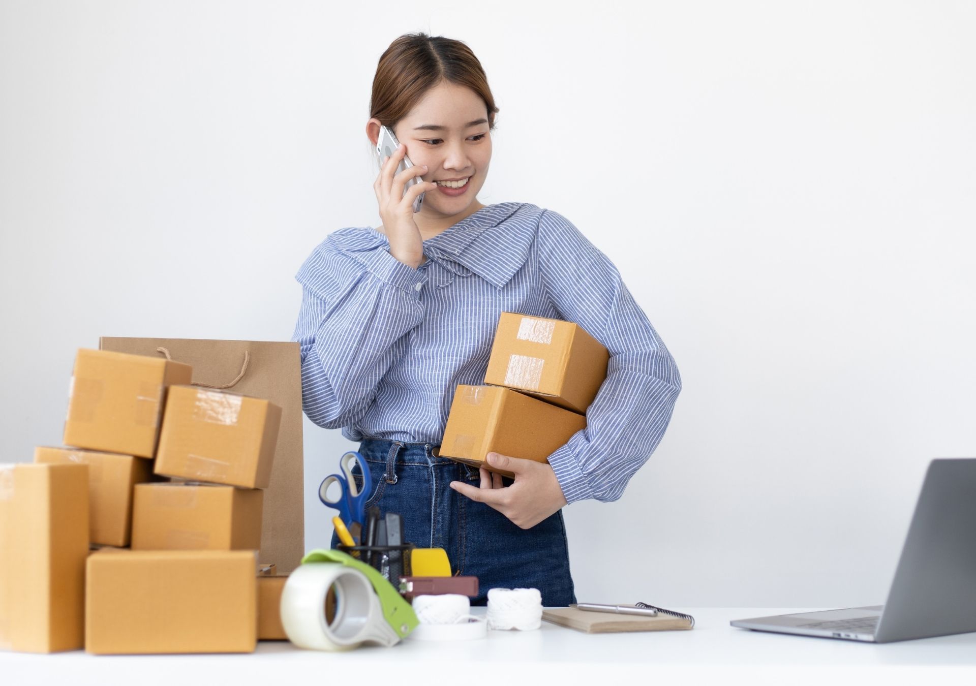 Woman on phone smiles, holding packages next to more boxes and a laptop on a white table.